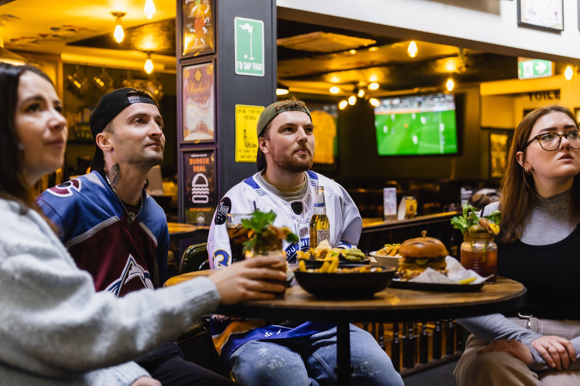 Friends in hockey jerseys watching a game over burgers and fries at The Locker Room